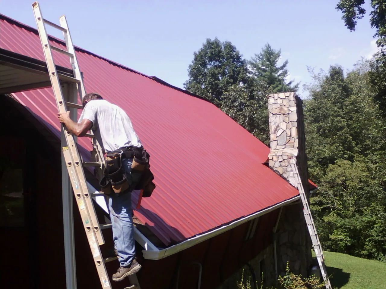 Roofer on a ladder beside a red metal roof and stone chimney — Soffit & Fascia Repair by Roofing Brothers in Rifle